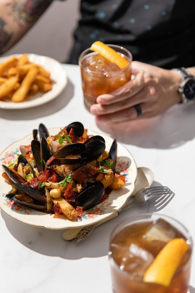 a dining table with a bowl of mussels and a man holding a cocktail as an example of cookbook photography